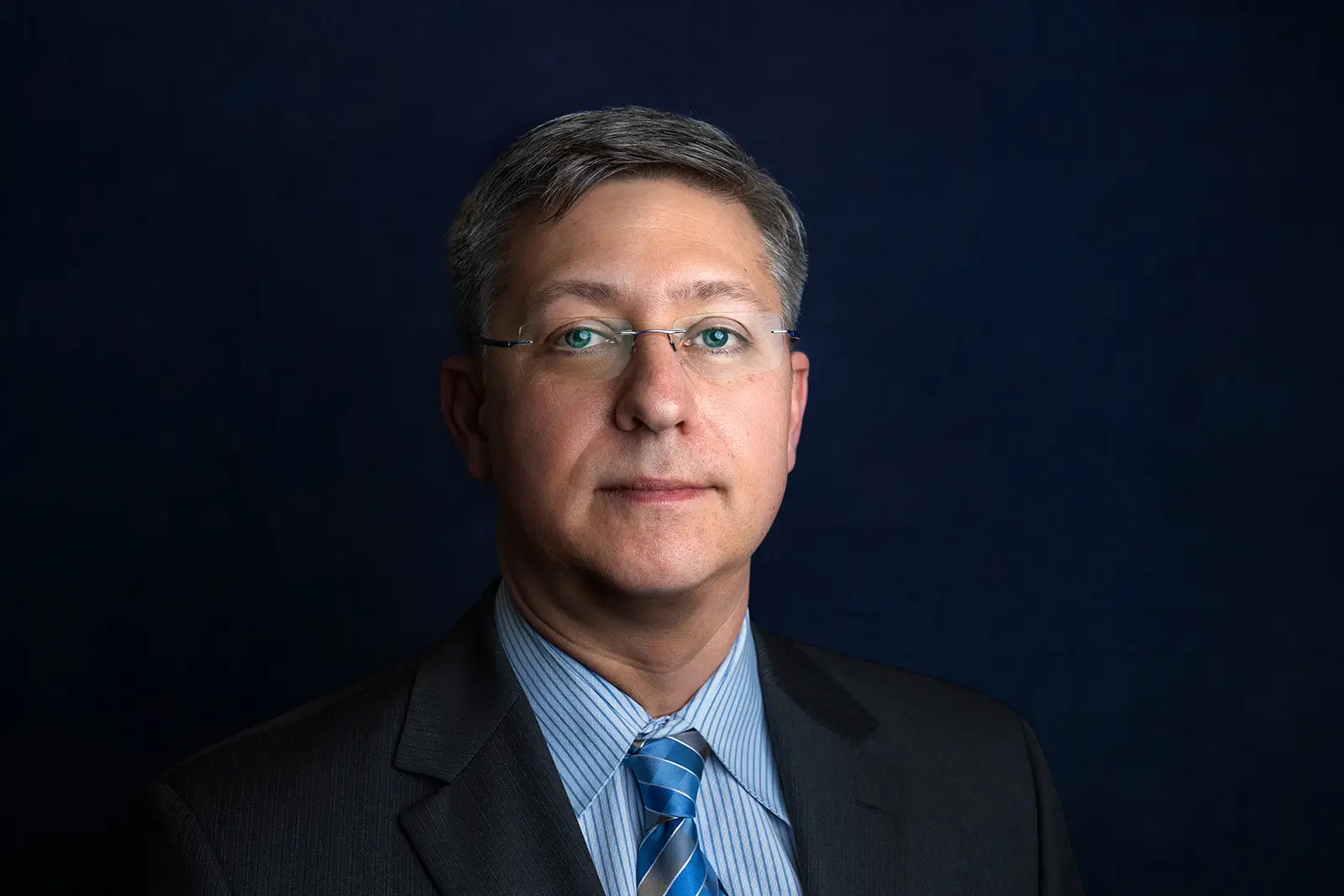 Studio headshot of a man in glasses and a striped shirt with blue tie against a deep navy background