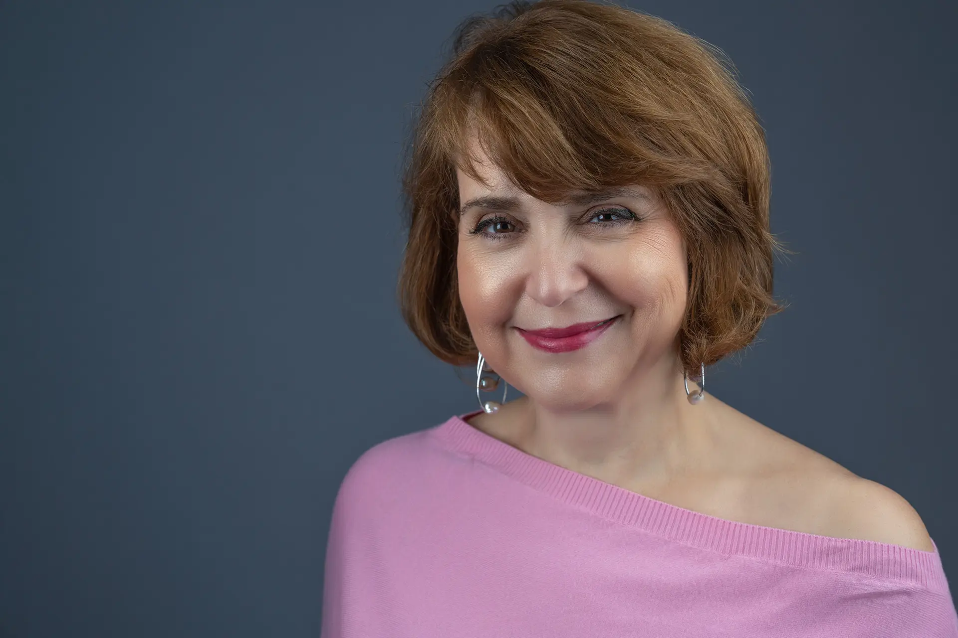 Woman with a brown bob haircut in a pink off-shoulder top smiling for a headshot against a dark blue-grey backdrop