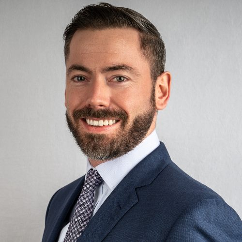 Bearded man in a navy suit and patterned tie smiling for a corporate headshot against a light grey backdrop