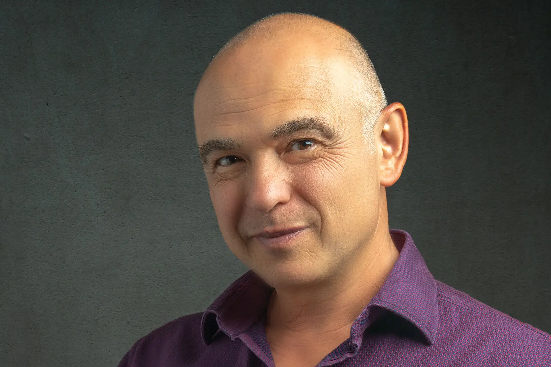 Bald man in a plum button-down shirt looking off-camera in a moody grey-toned studio portrait