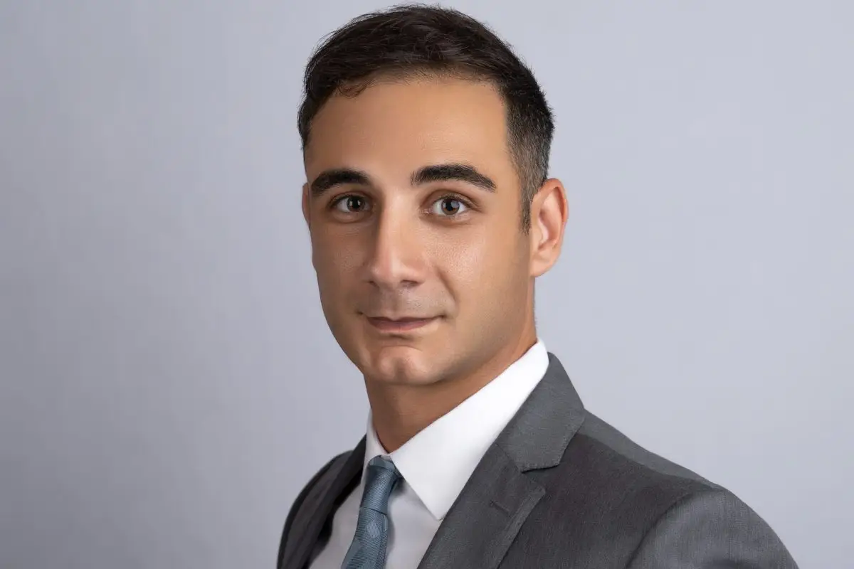 Man in a charcoal suit and light blue tie photographed at a three-quarter angle against a soft grey backdrop