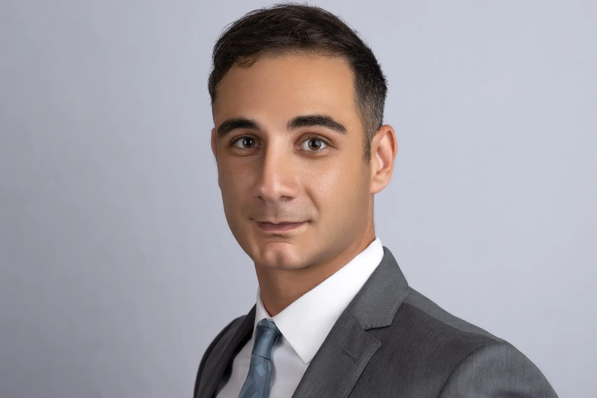 Man in a charcoal suit and light blue tie photographed at a three-quarter angle against a soft grey studio backdrop
