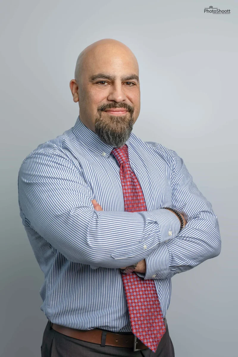 Bald man with a beard in a blue pinstripe shirt and red tie, arms crossed, corporate headshot on light grey